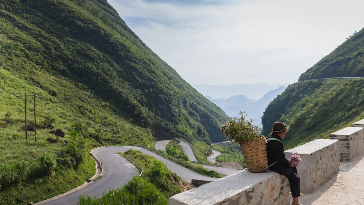 Woman with a basket on a wall above the hairpins of a pass road in Ha Giang