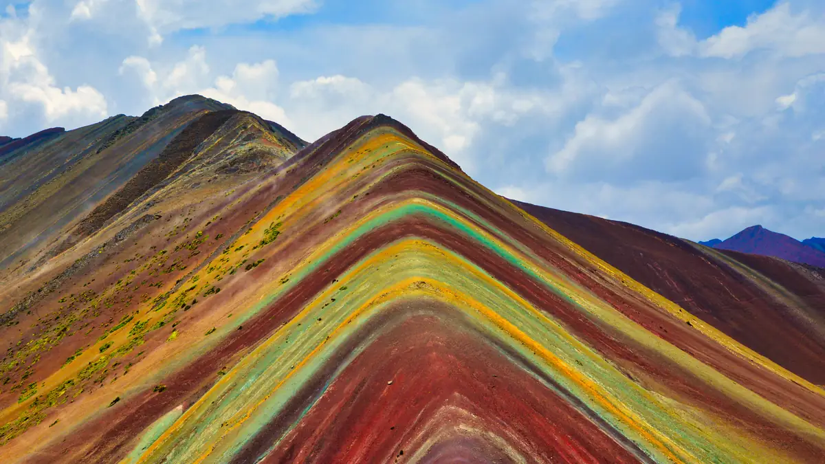 Der Vinicunca, die Regenbogenberg genannte Flanke aus roten, gelben und türkisen Schichten, unter blauem Himmel