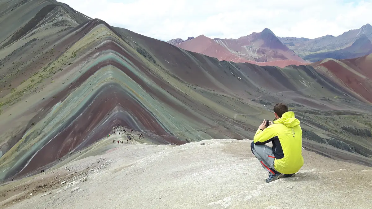 Ein Wanderer in gelber Jacke fotografiert vom Grat aus die Schichten des Vinicunca